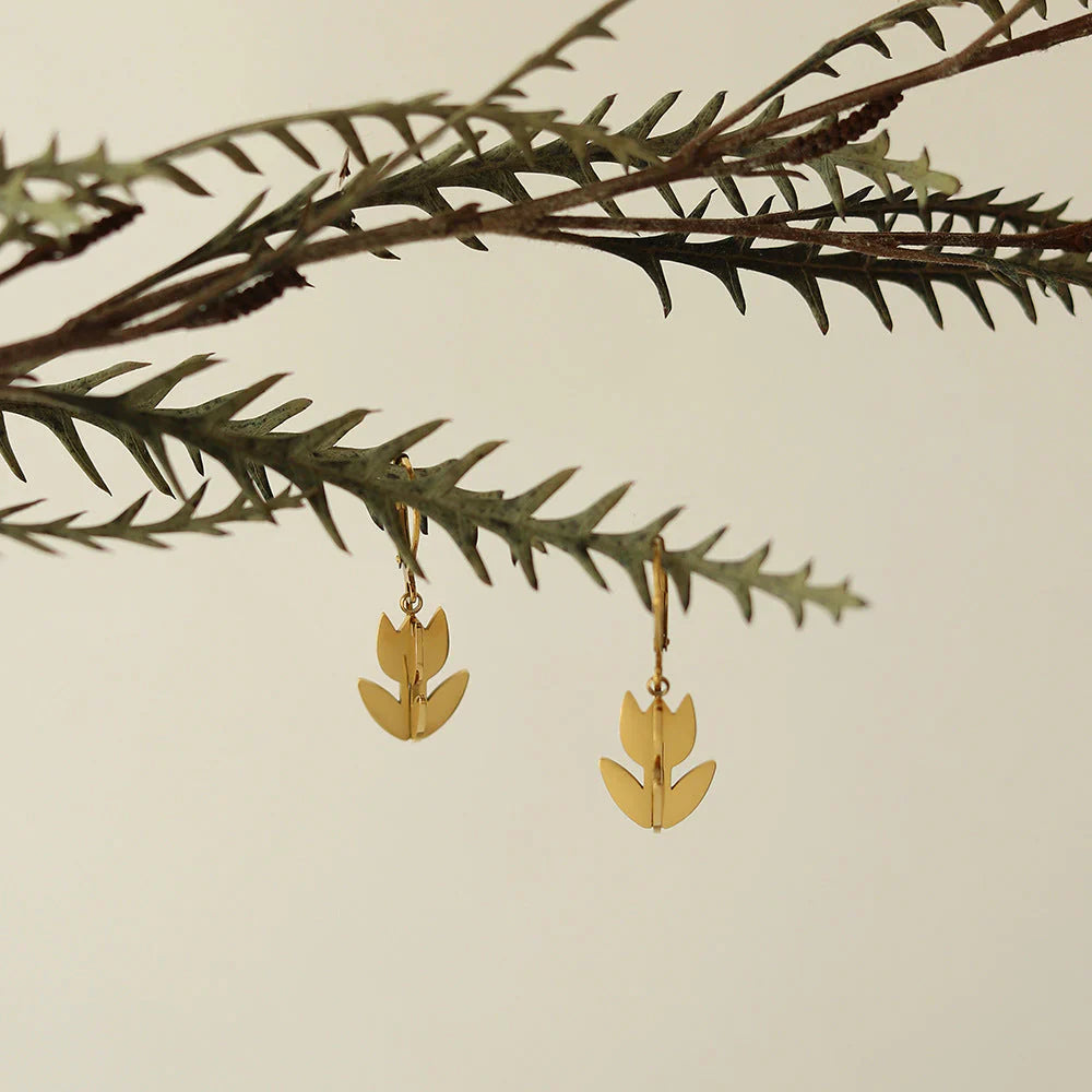 Gold leaf-shaped earrings hanging from a branch with a beige background